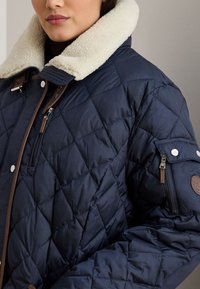 Σκούρο μπλε καπιτονέ μπουφάν με γιακά από μπεζ shearling, με καφέ δερμάτινες λεπτομέρειες, ασημένια κουμπιά και τσέπη με φερμουάρ στο μανίκι.