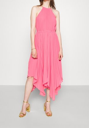 Žena v růžových bezrukávových plisovaných midi šatech s asymetrickým lemem a zlatými sandály na vysokém podpatku stojí před jednobarevným pozadím.