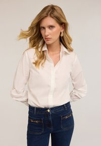 Camicia bianca con colletto strutturato, caratterizzata da dettagli di cucitura sottili. Abbinata a jeans in denim scuro con accenti dorati.