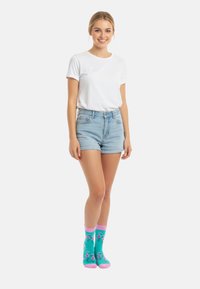 Türkise Socken mit pinken Akzenten, die Flamingo-Muster aufweisen; kombiniert mit einem weißen T-Shirt mit kurzen Ärmeln und Jeansshorts.