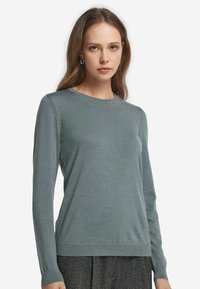 Kvinna klädd i en figurnära, långärmad, dämpad teal färgad rundhalsad tröja med mörkgrå, texturerade byxor, mot en enkel vit bakgrund.