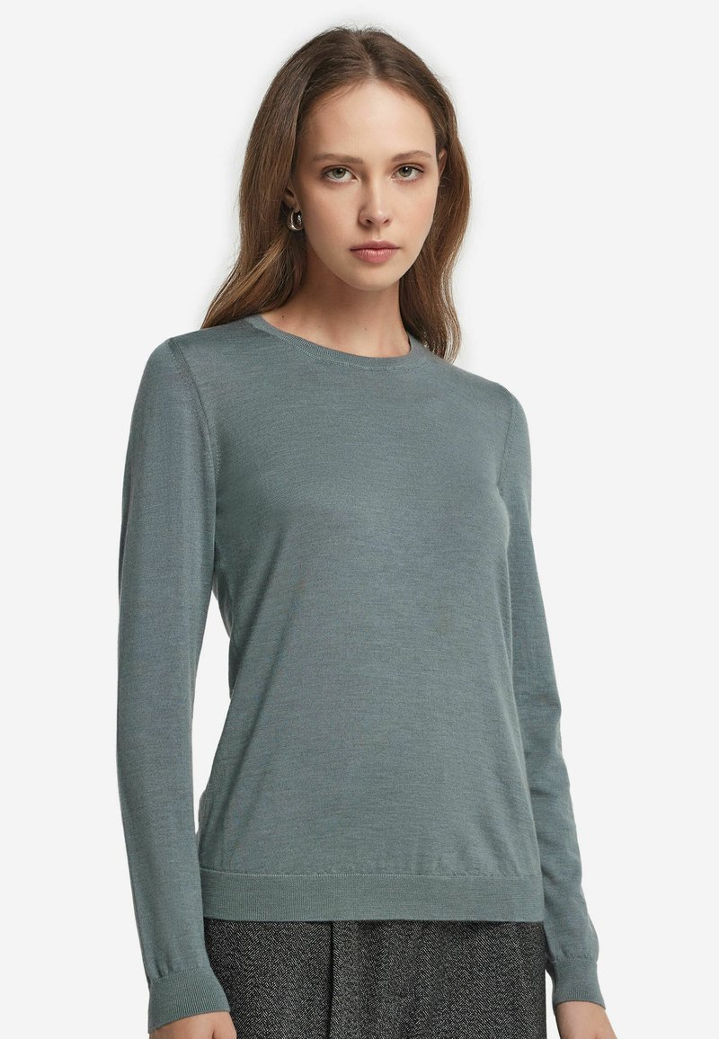 Kvinna klädd i en figurnära, långärmad, dämpad teal färgad rundhalsad tröja med mörkgrå, texturerade byxor, mot en enkel vit bakgrund.