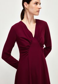 Burgundy pitkähiha dressi, jossa on V-pääntie, ja joka sisältää rypistettyä kangasta rinnassa ja soljuvan siluetin. Pehmeä ja venyvä materiaali.