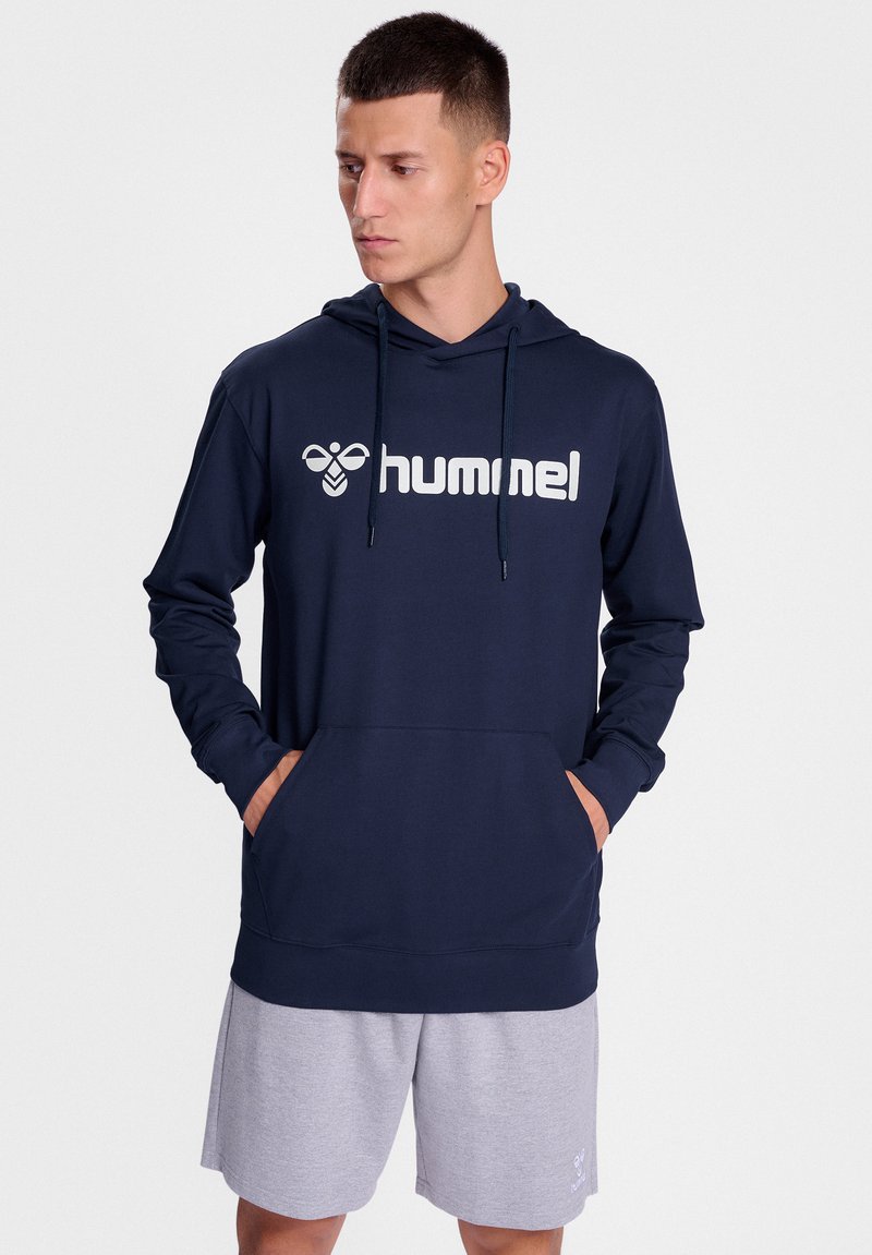 Marinblå hooded sweatshirt med logga, framficka i känguru-stil och dragsnoddar. Stylad med grå shorts som har en avslappnad passform och mjuk textur.