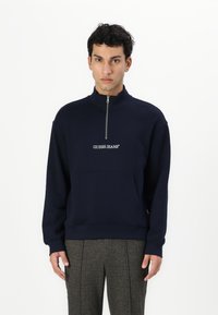 Mørkeblå sweatshirt med halv lynlås og broderet "GUESS JEANS" logo. Har en frontlomme og ribstrikkede manchetter for struktur.