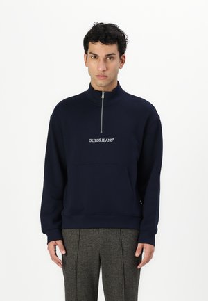 Navy blue sweatshirt with a half-zip collar and embroidered "GUESS JEANS" logo. Features a front pocket and ribbed cuffs for texture.
