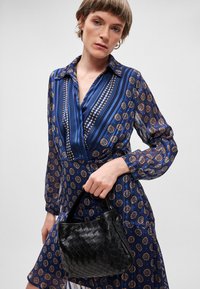 Juodos audinės rankinės su apvalia forma ir trumpa rankena, laikomos asmens, vilkinčio mėlyną raštuotą suknelę su ilgomis rankovėmis ir apykakle.