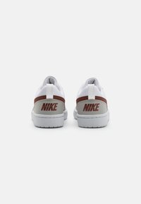 Witte sportieve schoenen met een rood en grijs ontwerp, voorzien van een opvallend Nike-logo op de hiel, een gestructureerd paneel en een platte witte tussenzool.