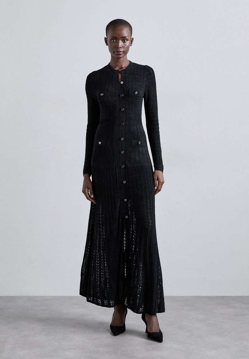 Sort strikket maxikjole med knaplukning foran, lange ærmer og to brystlommer; har ribbet struktur og en tætsiddende silhuet.