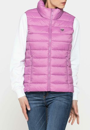 Doudoune rose avec un col montant, design matelassé horizontal, zip sur le devant et poches latérales, portée par-dessus une chemise à manches longues blanche.