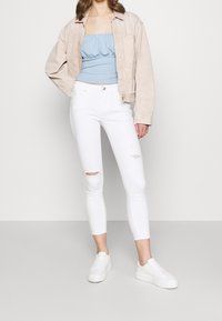 Žena v béžové bundě, světle modrém svršku s rouchou, bílých potrhaných skinny džínách a bílých platformových teniskách stojící na bílém pozadí.