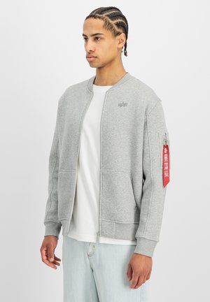 Joven con el pelo trenzado, vestido con una chaqueta gris clara con cremallera y una etiqueta roja en la manga, sobre una camisa blanca y unos vaqueros azul claro, mirando de lado.