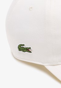 Gorra de béisbol blanca con visera curvada, que presenta un logo de cocodrilo bordado en verde en el costado. Fabricada con tejido texturizado.