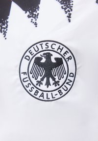 Έμβλημα μαύρου αετού με το κείμενο "Deutscher Fussball-Bund" πάνω σε λευκό ύφασμα, μέρος του σχεδίου ενός αθλητικού μπλουζιού.