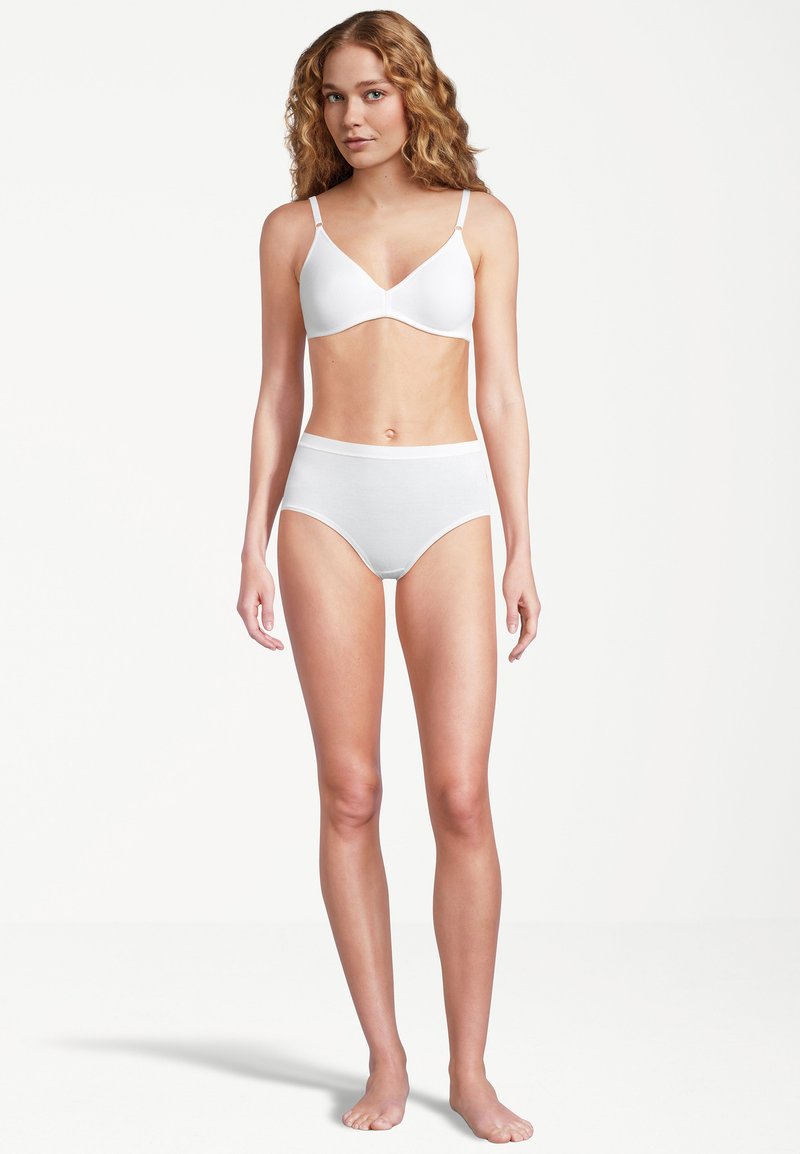 Femme aux cheveux blonds ondulés se tient pieds nus, portant un soutien-gorge blanc et des sous-vêtements taille haute, devant un fond blanc uni.