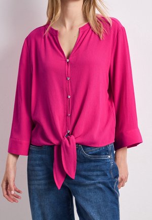 Donna che indossa una camicetta rosa acceso a maniche lunghe con bottoni anteriori annodata, abbinata a jeans di denim blu.