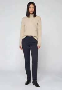 Beige kabelgebreide trui met hoge kraag en geribbelde textuur, gecombineerd met marineblauwe slim-fit broek en zwarte enkellaarsjes. Eenvoudig, strak design.