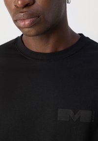 Černé bavlněné tričko s kulatým výstřihem, na hrudi s jemným, texturovaným logem v tmavě šedé barvě. Hladká, jednobarevná tkanina.