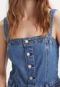 Jeanskjol i ljusblått med breda axelband, fyrkantig halslinje, fem silverknappar och en figursydd livdel med subtil sömnad.
