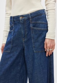 Τζιν παντελόνι σε σκούρο μπλε με φαρδιά γραμμή, διαθέτει ψηλή μέση, εμπρόσθιες τσέπες και μπρονζέ κουμπιά. Λείου υφής.