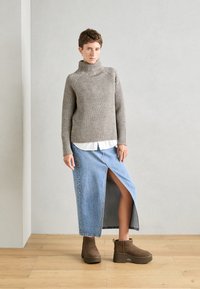Suéter beige grueso con canalé, camisa blanca debajo, falda de mezclilla larga con una abertura lateral, y botas marrones de tobillo con suelas gruesas.