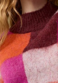 Bunter Strickpullover mit hohem, geripptem Kragen. Blöcke in Rosa, Orange und Burgunderrot bilden ein Patchwork-Muster. Weiches, strukturiertes Material.