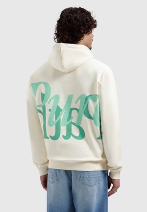 Man met krullend haar, draagt een crèmekleurige hoodie met grote blauwgroene gestileerde tekst op de achterkant en lichtblauwe spijkerbroek, van achteren gezien, lichte achtergrond.