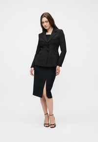 Γυναίκα που φοράει ένα εφαρμοστό μαύρο σακάκι με δομημένες ραφές και μαύρη μίντι φούστα με άνοιγμα μπροστά, συνδυασμένα με μαύρα σανδάλια με λουράκια.