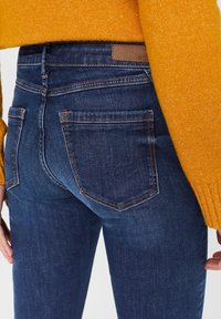 Skinny jeans in dark blue denim with orange stitching and two back pockets. The fabric has a smooth texture and slight sheen.