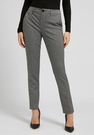 Vrouw draagt zwart-witte, nauwsluitende geruite broek gecombineerd met een zwarte top met lange mouwen en zwarte spitse hoge hakken.