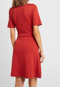 Žena oblečená v krátkých rukávech, šatách až ke kolenům v rezavě červené barvě, stojící zády k fotoaparátu na jednoduchém pozadí.