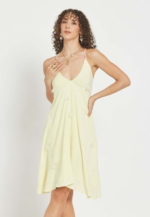 Mujer con cabello rizado que lleva un vestido sin mangas de color amarillo pálido con tirantes finos, complementado con pendientes, collar y anillo.