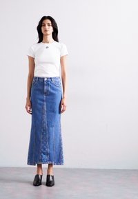 White t-shirt with round logo, paired with a long blue denim skirt featuring front pockets and visible stitching, and black slip-on shoes.