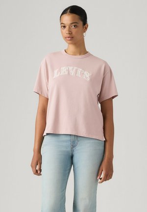 Joven con cabello oscuro recogido, llevando una camiseta Levi's rosa claro y vaqueros azul claro, de pie frente a un fondo liso.