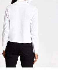 Eine Frau trägt ein weißes Langarmoberteil und schwarze Jeans, von hinten vor einem schlichten weißen Hintergrund gezeigt.