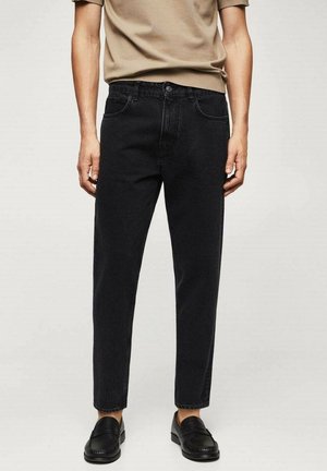 Homme portant un jean droit noir, une chemise beige à manches courtes et des mocassins noirs, debout devant un fond clair uni.