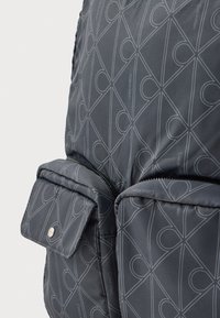 Mochila acolchada gris con patrón lineal, que cuenta con un compartimento con cremallera y un pequeño bolsillo con solapa y cierre de presión.