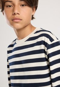 Junge Person mit kurzem braunem Haar, die ein navyblau-weiß gestreiftes Langarmshirt gegen einen schlichten Hintergrund trägt.