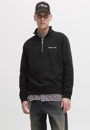 Giovane uomo con felpa nera mezza zip con logo "Originals Studio", camicia a quadri sotto, cappello nero con scritta bianca e jeans neri sbiaditi.