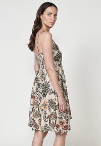 Virágmintás, ujj nélküli ruha könnyű anyagból; smocked bodice-al rendelkezik, krém és színes virágokkal és lombozattal a háttérben.