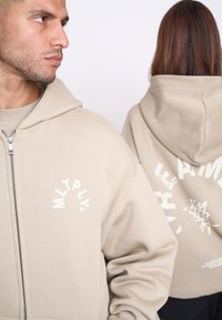 Beige zip-hoodie med frontlogo og stort grafik på ryggen. Blødt stof, afslappet pasform og en hætte for ekstra dækning.