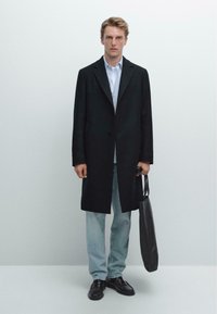 Mann trägt einen schwarzen Überrock, helle Jeans und schwarze Schuhe, hält eine schwarze Tragetasche vor einem schlichten weißen Hintergrund.