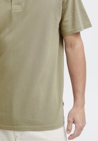 Oliven grønn polo skjorte med teksturert stoff, korte ermer og krage. Sidesøm med et lite logo-merke nederst.