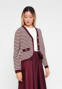 Γυναίκα με μπορντό και λευκές ρίγες κάρντιγκ με ασημένια κουμπιά πάνω από λευκή μπλούζα και μπορντό φούστα, κοιτάζοντας προς τα κάτω αριστερά.