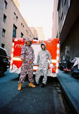 Two individuals wear matching hooded jumpsuits; one pink with black patterns, the other light gray. They stand against a red emergency vehicle.