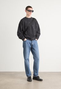 Sudadera negra con acabado texturizado, jeans azules, zapatos negros y gafas de sol de montura cuadrada. El atuendo incluye una correa cruzada negra.