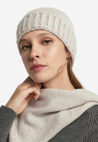 Dziergana czapka w jasnobeżowym kolorze z pionowym prążkowaniem i srebrnymi koralikami, zestawiona z miękkim, lekkim szalem ozdobionym rozsianymi dekoracjami.