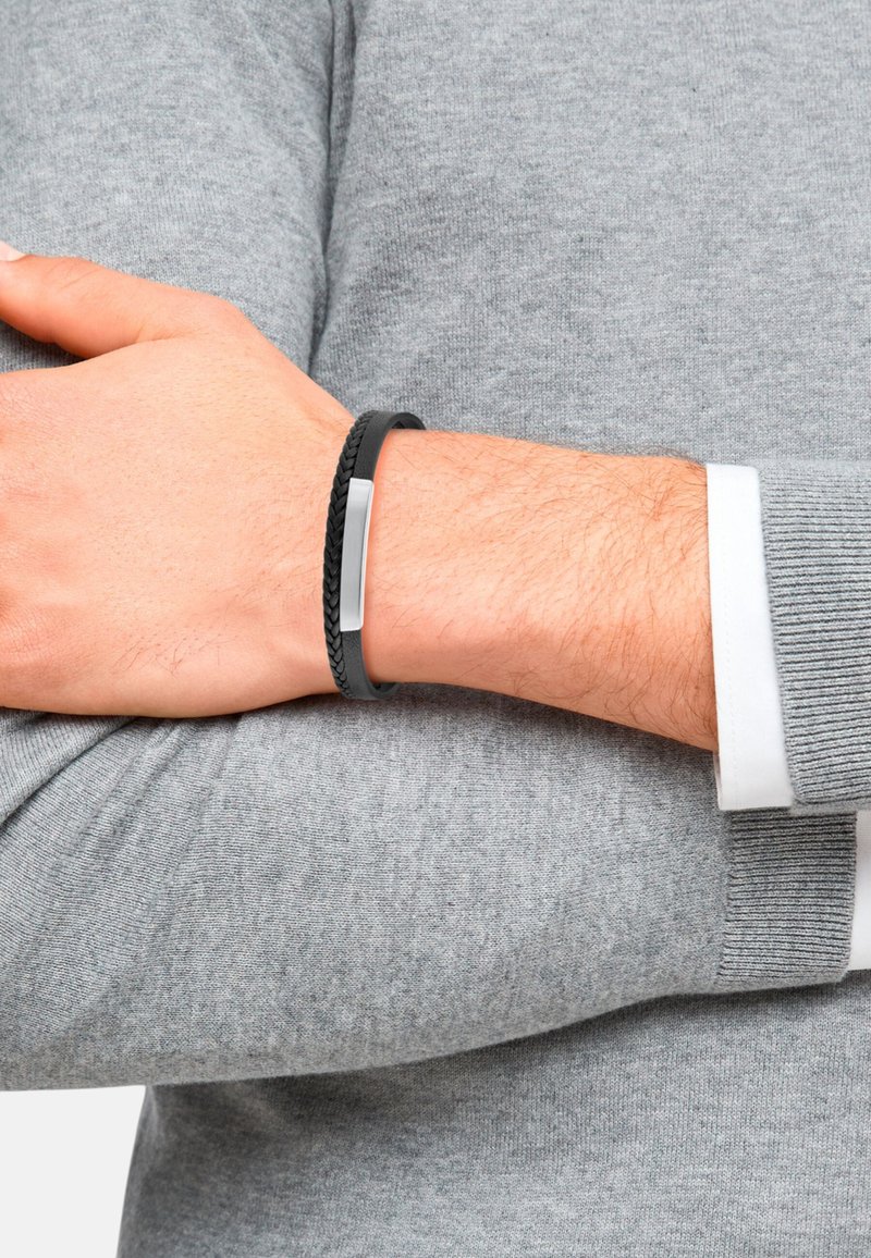 Pulsera trenzada negra con un acento plano de plata, llevada en una muñeca, contra un fondo de suéter gris texturizado. Materiales suaves y duraderos.