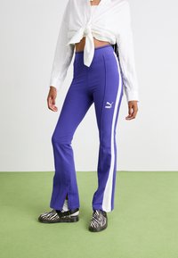 Lila utsvängda byxor med vita sidostripes, tillverkade av mjukt tyg, med en Puma-logotyp. Kombinerade med svart och vit zebramönstrade skor.