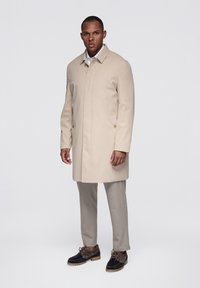 Abrigo beige hasta la rodilla, hecho de material liso, con cierre de botones ocultos y bolsillos laterales. Combinado con pantalones gris claro y zapatos negros.